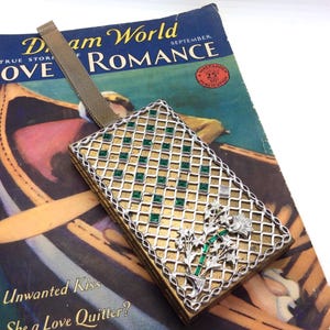 May include: A vintage compact mirror with a gold and silver geometric design, featuring green squares and floral embellishments. The mirror is placed on a vintage magazine with the title "Dream World Romance".