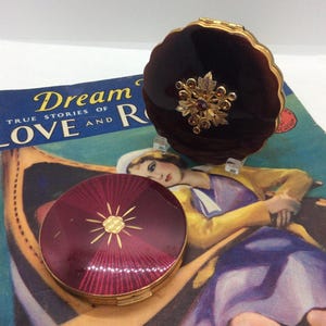Op de afbeelding: Twee vintage compactpoeders. Eén is diep bordeauxrood met een gouden zonnestraalontwerp. De andere is een donkerbruin compactpoeder met geschulpte rand en gouden bloemversiering. De achtergrond bevat een vintage magazinecover.