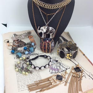 Könnte beinhalten: Eine Sammlung von Vintage-Schmuck, darunter Halsketten, Armbänder und Ohrringe. Die Halsketten sind gold- und silberfarben, mit einem Elefantenanhänger. Armbänder haben Perlen und Strasssteine. Ohrringe haben schwarz-goldene geometrische Designs. Der Schmuck wird auf einer Vintage-Magazinseite präsentiert.