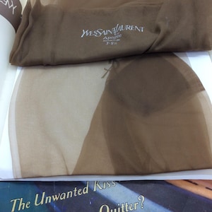 Puede incluir: Un par de medias transparentes de color marrón con el logotipo de Yves Saint Laurent y el texto Apogée 3-9 1/2 impreso en blanco sobre la tela. Las medias están dobladas y superpuestas, mostrando un material ligero y translúcido.