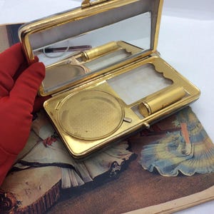 May include: Open gold-toned compact with a mirror, lipstick, and a powder compartment. The compact is rectangular with rounded corners and a clasp. The interior features a mirror, lipstick, and a powder compartment. The compact is open, revealing the contents.