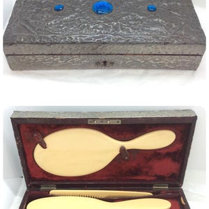 Puede incluir: Antiguo juego de tocador en una caja de metal texturizado con detalles de vidrio azul. La caja se abre para revelar un interior de terciopelo rojo con cepillos, un peine y un espejo de color marfil. Un kit de aseo vintage.