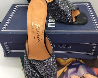 Pode incluir: Mules pretas de bico aberto com glitter preto e biqueira de verniz. Os sapatos estão em uma caixa azul marinho. A palmilha tem a marca "Monia" e "Made in Italy".