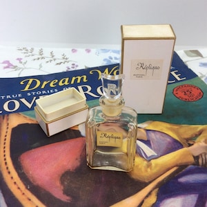 May include: A vintage perfume bottle labeled "Réplique" by Raphael Paris, with a clear glass bottle and a decorative stopper. The bottle is accompanied by its original box and a small, open container. The background features a vintage magazine.