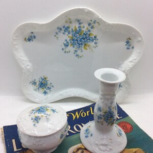 May include: White porcelain serving tray, trinket box, and candlestick holder, all adorned with blue floral patterns. The tray has a unique, scalloped shape. The items are arranged on a vintage book.