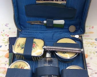 May include: A blue zippered travel case containing a grooming set with a hairbrush, three round containers, and a glass bottle. Each item features a cream-colored top with a floral pattern and silver-tone trim. A small knife is secured in the lid.