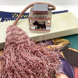 May include: A clear glass perfume bottle with a silver-coloured top and a brown and black Scottie dog design. A pink tassel and a book are in the background.