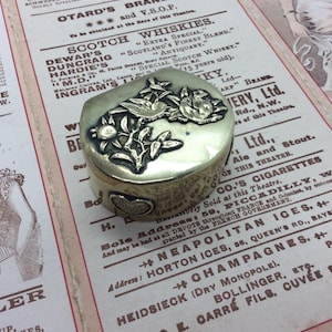 May include: An antique brass box with a hinged lid, featuring detailed floral and bird designs. The box is rectangular with rounded edges, set against a vintage paper background with faded text. Likely used for jewellery or small keepsakes, it is a charming vintage item.