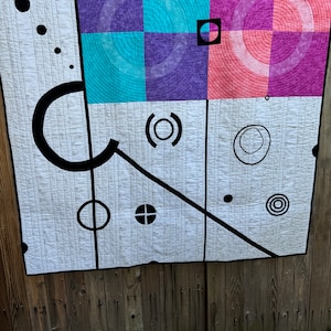 以下が含まれることがあります： 黒い幾何学模様の白いキルト。キルトは4つの正方形で構成されており、それぞれ異なる色の背景になっています。正方形は紫、ピンク、青、白です。黒いデザインには、円、線、十字が含まれています。