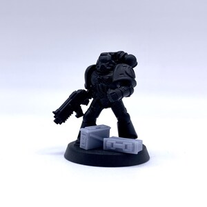 May include: A black plastic miniature of a space marine holding a gun and standing on a black base with two gray plastic ammo boxes.