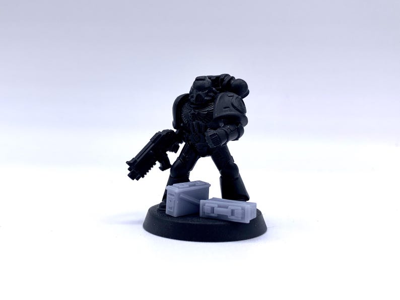 May include: A black plastic miniature of a space marine holding a gun and standing on a black base with two gray plastic ammo boxes.