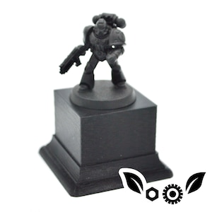 Puede incluir: Una figura en miniatura negra de un marine espacial de pie sobre una base cuadrada negra. La figura sostiene un arma en una mano y una espada en la otra.