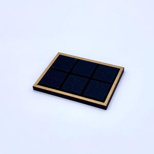 May include: A square wooden coaster with a gold-colored frame and six black square inserts. The coaster is set against a white background. The design is simple and modern.