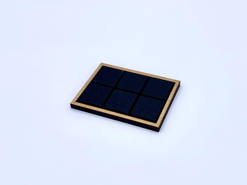 May include: A square wooden coaster with a gold-colored frame and six black square inserts. The coaster is set against a white background. The design is simple and modern.