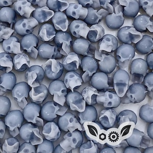 Op de afbeelding: Een verzameling kleine, lichtblauwe kralen in de vorm van een schedel. De kralen hebben een gedetailleerd, driedimensionaal ontwerp met zichtbare oogkassen, neusholtes en tanden. Een witte sticker met een zwarte afbeelding staat rechtsonder.