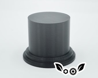 May include: A black, cylindrical pedestal with a smooth, rounded top and a slightly flared base. The pedestal is made of a textured material, possibly plastic or resin.
