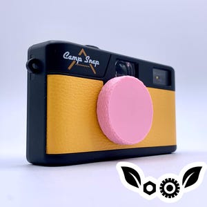 May include: A black and yellow camera with a pink lens cover. The camera has the words "Camp Snap" printed on the top. The camera has a retro design and is likely a film camera.