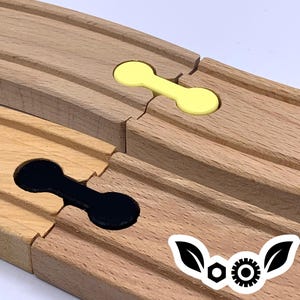 Pode incluir: Duas peças de trilho de trem de madeira com um conector preto e amarelo. O conector é um design simples com dois círculos conectados por uma linha. As peças de trilho são de madeira marrom claro.