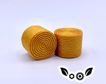 May include: Two miniature, cylindrical hay bales in shades of yellow and orange sit on a white background. The bales have a spiral design on the top and bottom, with a textured, woven appearance on the sides. A decorative logo is in the bottom right corner.