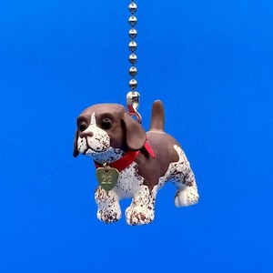 May include: A brown and white dog figurine with a red collar and a heart-shaped tag hanging from the collar. The figurine is attached to a silver chain.