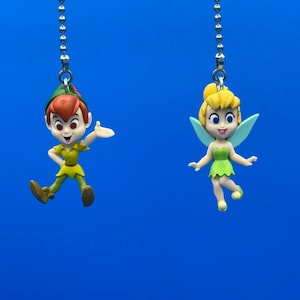 Op de afbeelding: Twee decoratieve trekkoorden voor plafondventilatoren met stripfiguren. Eén trekkoord toont Peter Pan in een groene outfit met een rode pet. De andere toont Tinkerbell in een groene jurk met blauwe vleugels. Beide hangen aan zilveren kettingen tegen een blauwe achtergrond.