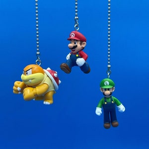 Może przedstawiać: Trzy wiszące figurki postaci z Mario: Mario, Luigi i Bowser. Figurki są przymocowane do srebrnych łańcuchów i wiszą na niebieskim tle.