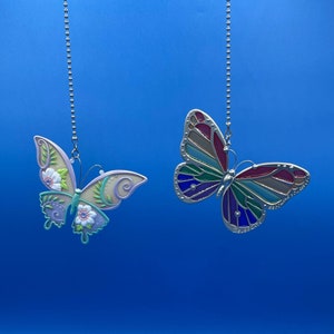 Peut inclure: Deux ornements de papillon décoratifs suspendus à des chaînes argentées sur un fond bleu. Le papillon de gauche est violet, vert et blanc avec des détails floraux. Le papillon de droite est rouge, vert, bleu et violet avec des détails ressemblant à du vitrail.