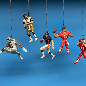 May include: Five football player ornaments hanging from chains. The ornaments are made of plastic and feature different players in various poses. The players are wearing uniforms in different colours, including red, white, blue, and black.