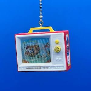 Op de afbeelding: Een rood en wit plastic speelgoedtelevisie met een trekkoord. De televisie heeft een gele knop en een afbeelding van een cartoon scène erin. De televisie hangt aan een zilveren ketting.