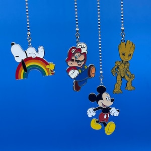 Peut inclure: Quatre breloques de personnages colorés suspendus à des chaînes argentées sur un fond bleu. Les breloques représentent Mickey Mouse, Mario, Groot et Snoopy sur un arc-en-ciel.