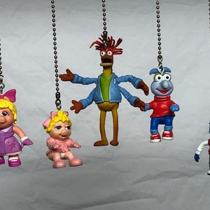 May include: Five Muppet character figurines hanging from silver chains. The characters are Miss Piggy in a pink dress, a pig in a pink dress, Animal in a blue jacket, a blue monster in a red shirt, and a blue monster in a red sweater.