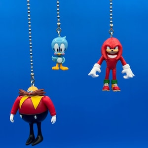 May include: Three character-themed ceiling fan pulls. The characters are from the Sonic the Hedgehog franchise. The characters are Dr. Eggman, Knuckles, and a blue bird. Each character is suspended from a silver ball chain against a blue background.