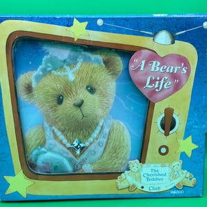 Puede incluir: Una caja azul y amarilla con una imagen de un oso de peluche que lleva un collar y una corona. La caja dice "A Bear's Life" y "The Cherished Teddies Club".