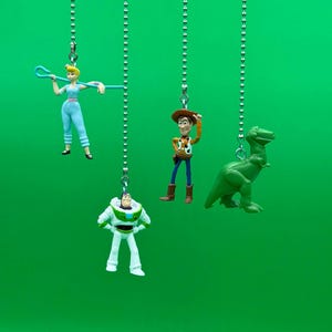 May include: Four Toy Story character figurines hanging from silver chains against a green background. The characters are Woody, Buzz Lightyear, Jessie, and the dinosaur Rex.