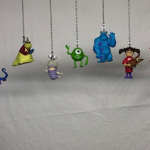 May include: Seven colorful plastic figurines hanging from silver chains. The characters include a purple monster, a green monster with one eye, a blue monster, a girl with red hair, a red monster, and a yellow monster with a red cape.