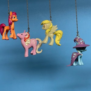 May include: Four colorful My Little Pony figurines hanging from silver chains. The ponies are pink, orange, yellow, and blue. The blue pony is wearing a purple hat with stars.