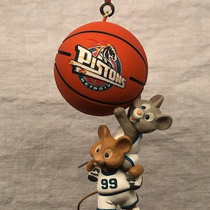May include: A basketball-themed ornament with a basketball bearing the Detroit Pistons logo, suspended from a chain. Two cartoon mice are shown, one wearing a jersey with the number 99, playing basketball. The ornament is made of a durable material.