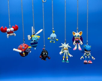 May include: A set of eight colourful character charms hanging from silver chains. The characters include a red crab, a ladybug, a blue bee, a blue and white character with a single eye, a white hedgehog, a blue and white character with a single eye, a pink and white bat-like character, and a blue and silver robot.