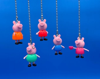 Könnte beinhalten: Fünf Peppa Wutz-Charms, die an silbernen Kugelkettchen vor blauem Hintergrund hängen. Die Charms sind rosa und tragen Outfits in Orange, Rot, Blau, Grün und Pink. Jeder Charm hat schwarze Schuhe.