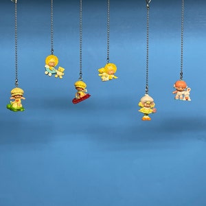 May include: Nine colourful hanging figurines of girls with different outfits and accessories. The figurines are attached to silver chains and are hanging against a blue background.