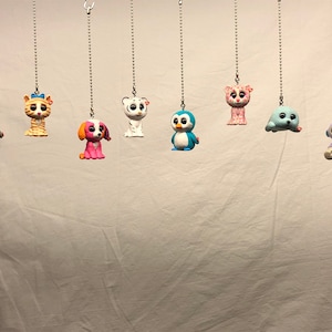 Op de afbeelding: Negen kleurrijke pluche speelgoedjes die aan zilveren kettingen hangen. De speelgoedjes omvatten een kat, een hond, een pinguïn, een aap, een zeehond en een uil.