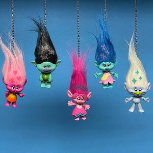 Puede incluir: Seis muñecos Troll coloridos colgando de cuerdas. Los muñecos tienen diferentes colores y estilos de cabello, incluyendo rosa, azul, blanco y negro. Cada muñeco tiene una expresión facial y características únicas.