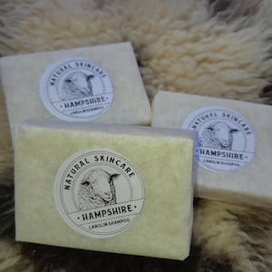 May include: Three rectangular bars of natural skincare lanolin shampoo, wrapped in off-white paper. Each bar features a circular sticker with a sheep illustration and the text "NATURAL SKINCARE HAMPSHIRE LANOLIN SHAMPOO". The background is a textured, light brown surface.