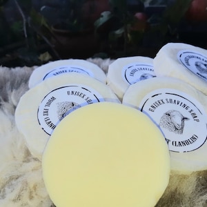 May include: Several round, off-white unisex shaving soaps with a pale yellow centre. Each soap is wrapped in white paper with a label featuring a sheep illustration and the text "Unisex Shaving Soap" and "Wool Fat (Lanolin)".