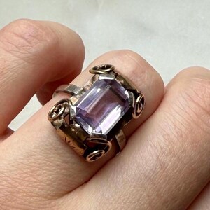 Op de afbeelding: Een ring met een rechthoekige, lichtpaarse gefacetteerde edelsteen in een metalen band. De zetting is voorzien van decoratieve, gekrulde metalen accenten op elk van de vier hoeken van de steen, wat een duidelijk kader vormt voor de centrale edelsteen.