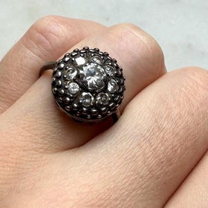 Op de afbeelding: Een ring van donker metaal met een ronde clusterzetting. Het midden is versierd met een enkele heldere ronde edelsteen, omringd door een ring van kleinere heldere stenen en een getextureerde, kralenachtige rand rondom het ontwerp.