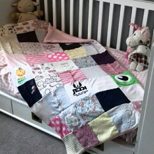 Pode incluir: Uma colcha de retalhos feita à mão em um berço, com quadrados de vários tecidos em rosa, branco e outras cores. A colcha inclui um patch "Jedi" e outros desenhos. Animais de pelúcia, incluindo um cordeiro e um unicórnio, estão na cama.