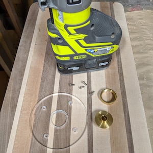May include: A yellow and black Ryobi router with a clear plastic base, a brass ring, and a brass plate on a wooden surface. The router is on top of the base and the ring and plate are below the base.