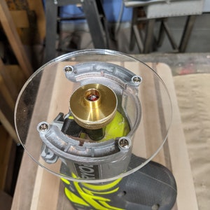 May include: A close-up view of a router bit attached to a router. The router bit is made of brass and has a small hole in the center. The router is attached to a clear plastic base with screws.
