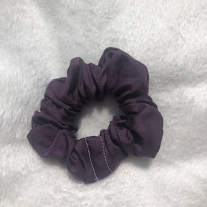 May include: A purple scrunchie made of soft fabric with a white stitch detail.